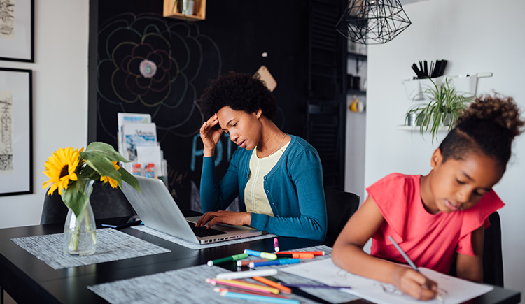 Women sitting on a laptop while looking stressed with a child coloring next to her