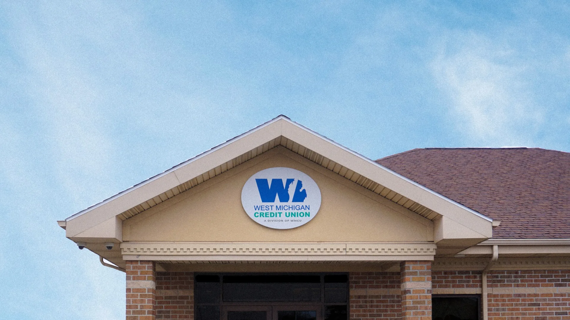 Front view of a brick building entrance with a round sign reading West Michigan Credit Union beneath a gabled roof on a clear day