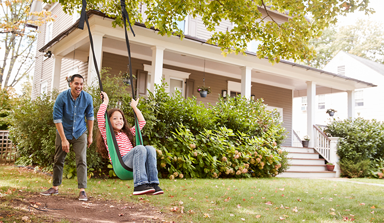 Older man pushing a young girl on a tree swing in front of a house during summer