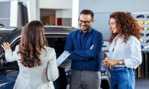 A car salesman is showing new cars to a couple. Car sales business. Manager talking to couple, showing them new auto at dealership shop. Young family selecting vehicle, looking at modern automobile at showroom