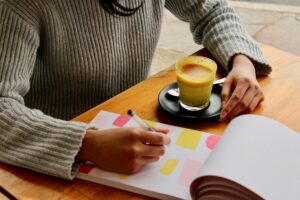 Woman in beige sweater writing in a journal at a table with a latte next to her hand.