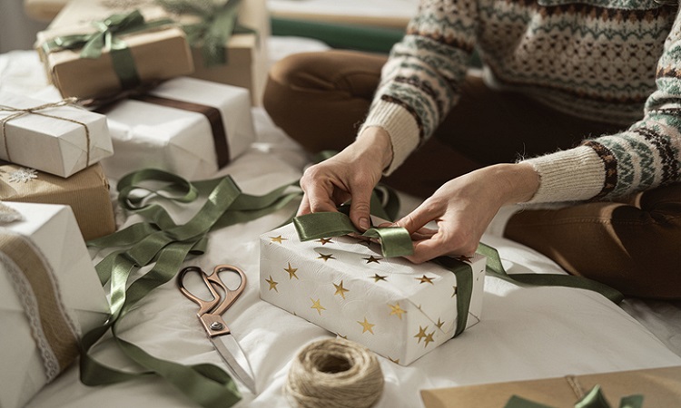 Hands of unrecognizable caucasian woman sitting on bed and packing Christmas gift at home