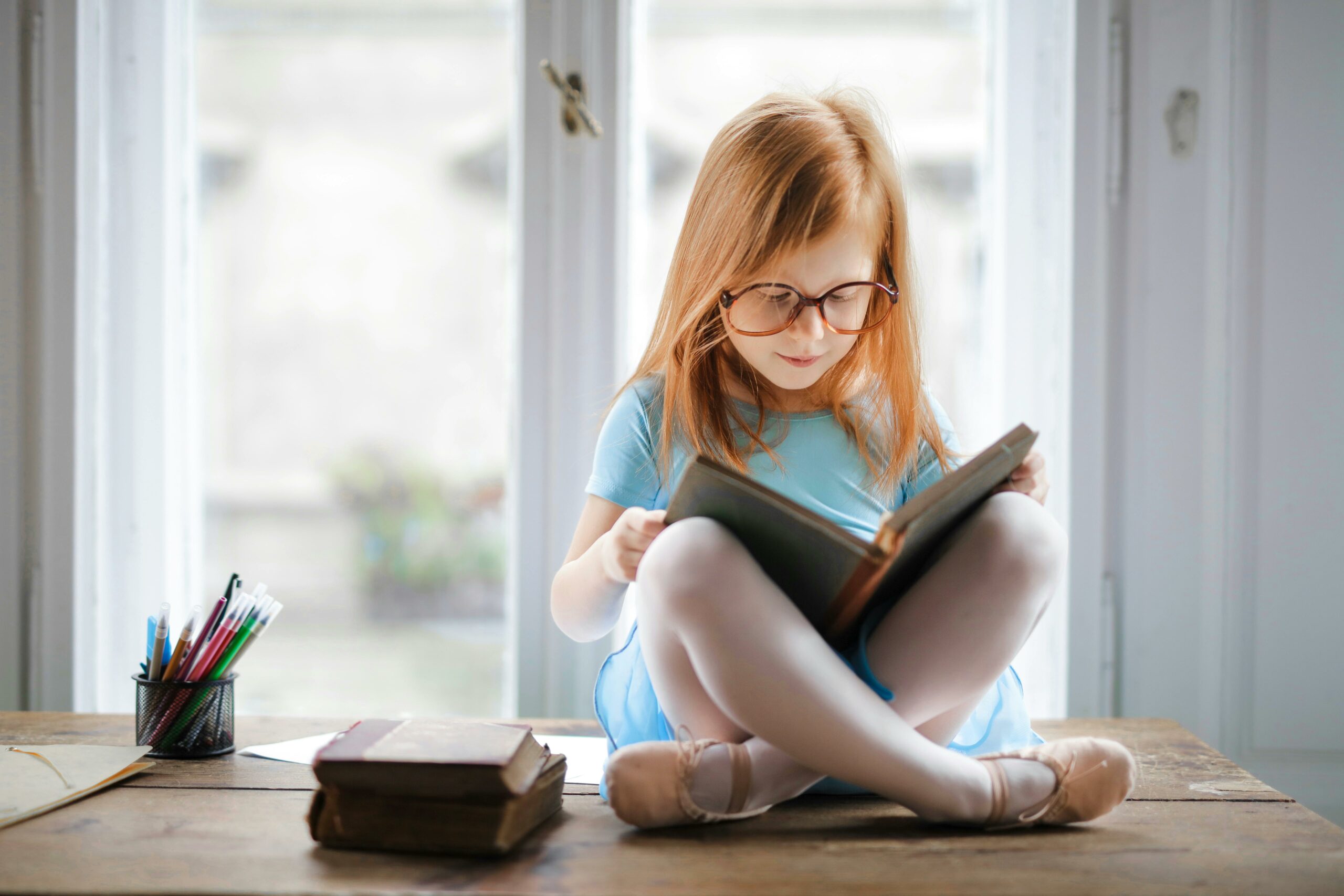 Young girl with red hair and glasses reads book on table.